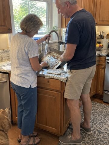 At a kitchen sink beneath a window in mid-afternoon, a couple in their sixties rinse a roasting pan together, one holding the tap while the other scrubs.