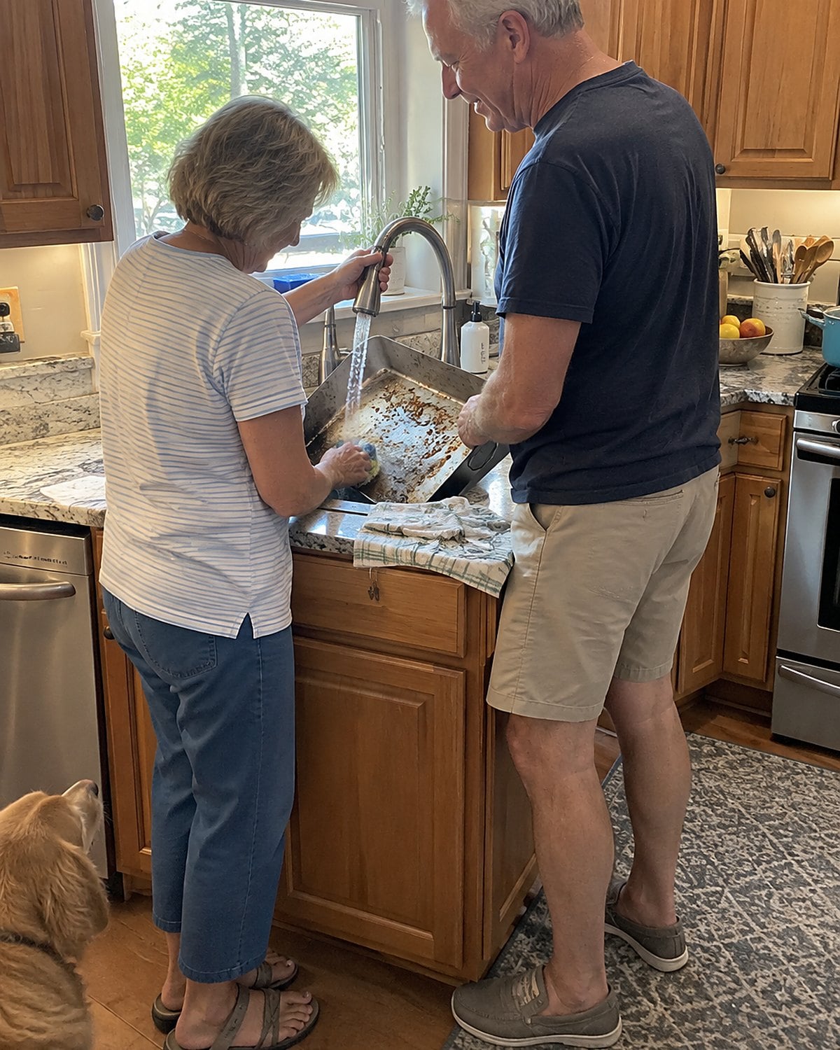 At a kitchen sink beneath a window in mid-afternoon, a couple in their sixties rinse a roasting pan together, one holding the tap while the other scrubs.