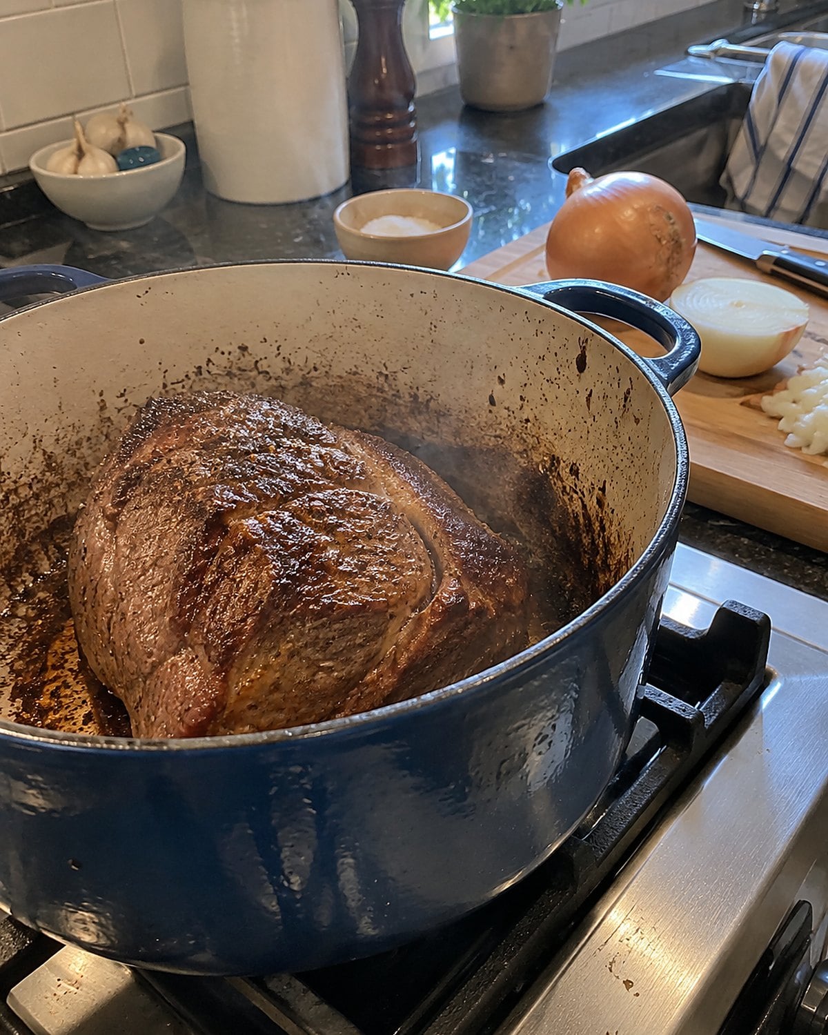 A roast browning in a heavy pot on the stove while an onion waits on the cutting board.