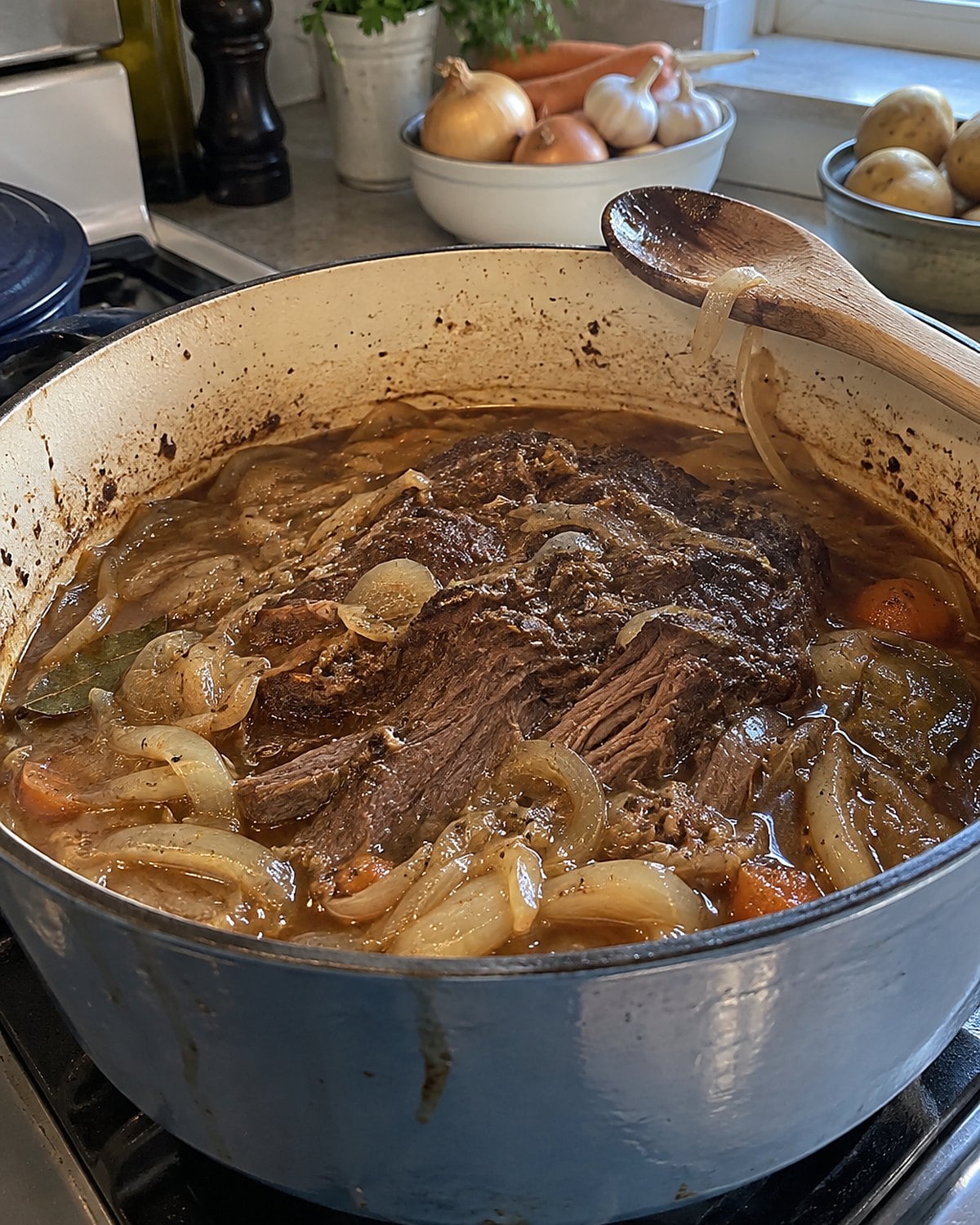 A Dutch oven of pot roast and onions with a wooden spoon resting against its rim.
