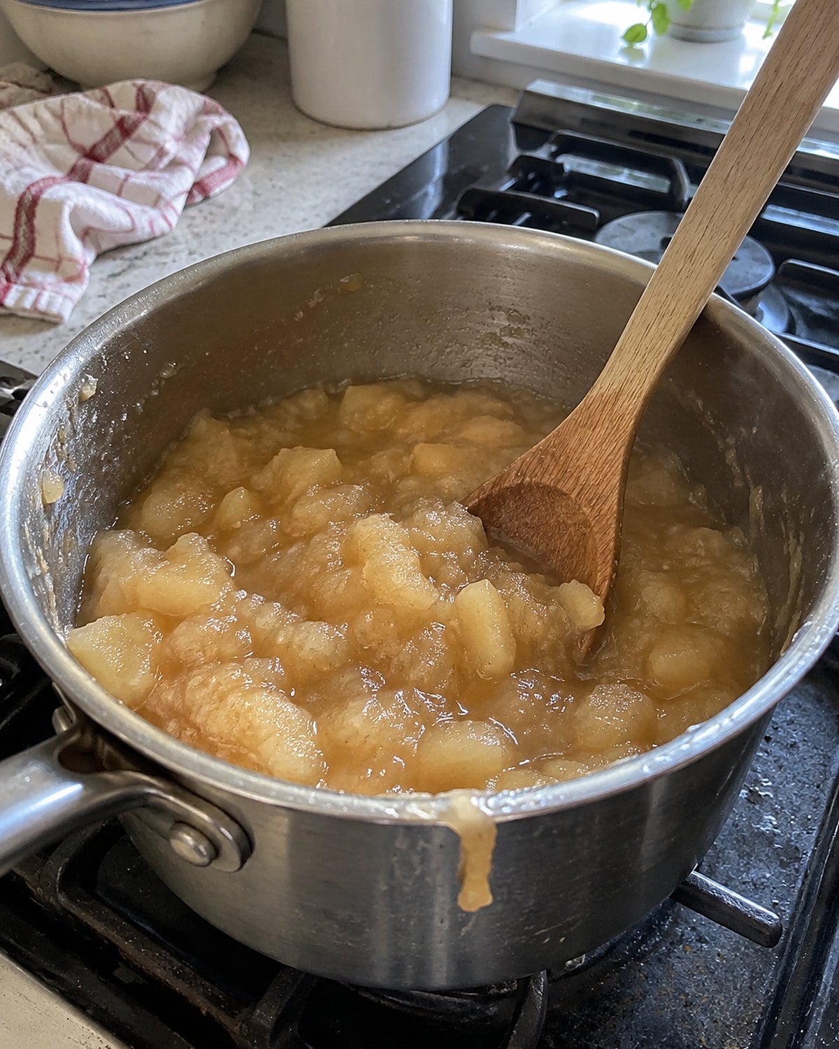A wooden spoon standing in a small saucepan of chunky applesauce.