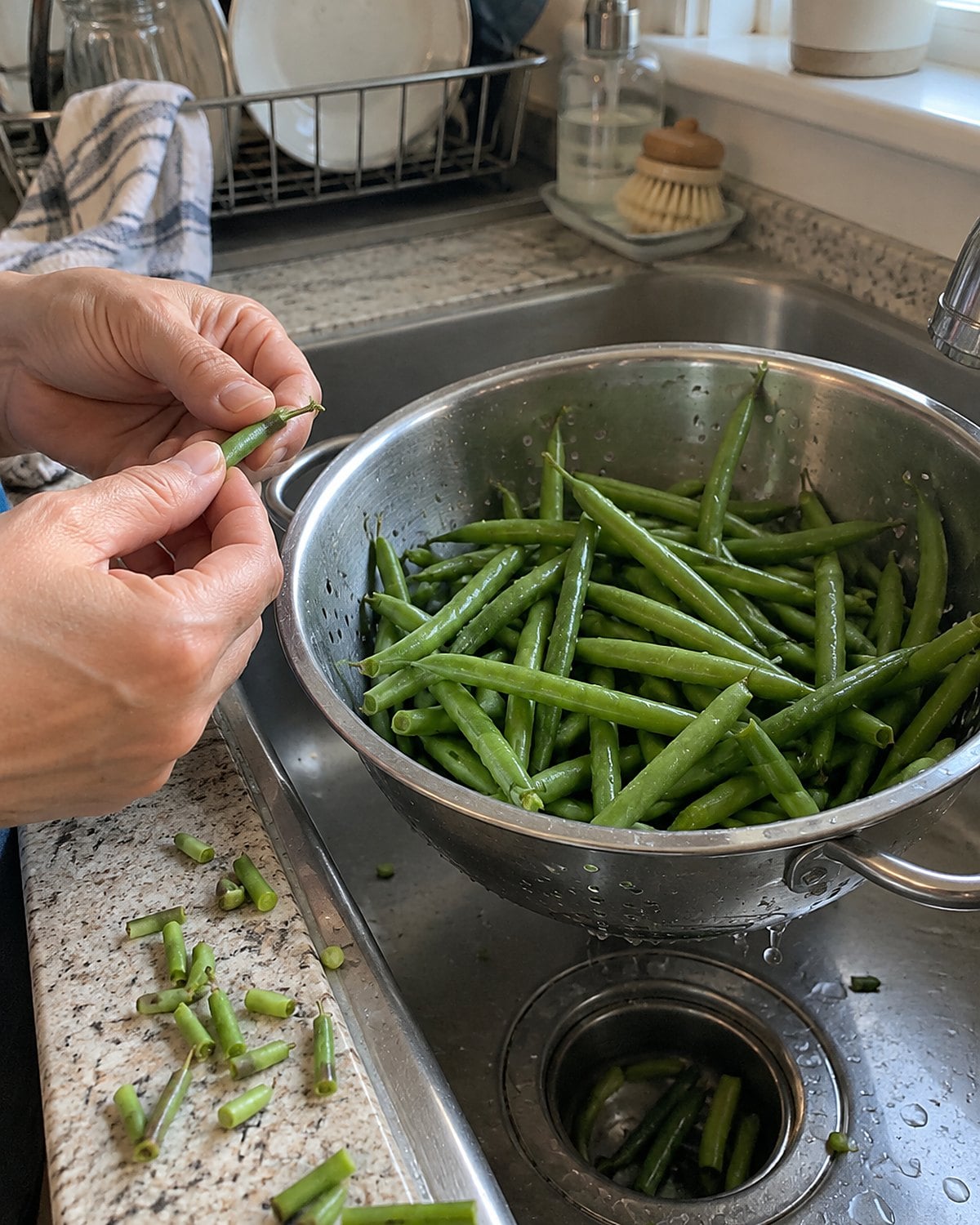 Fresh green beans dripping in a colander at the sink, with snapped ends nearby.