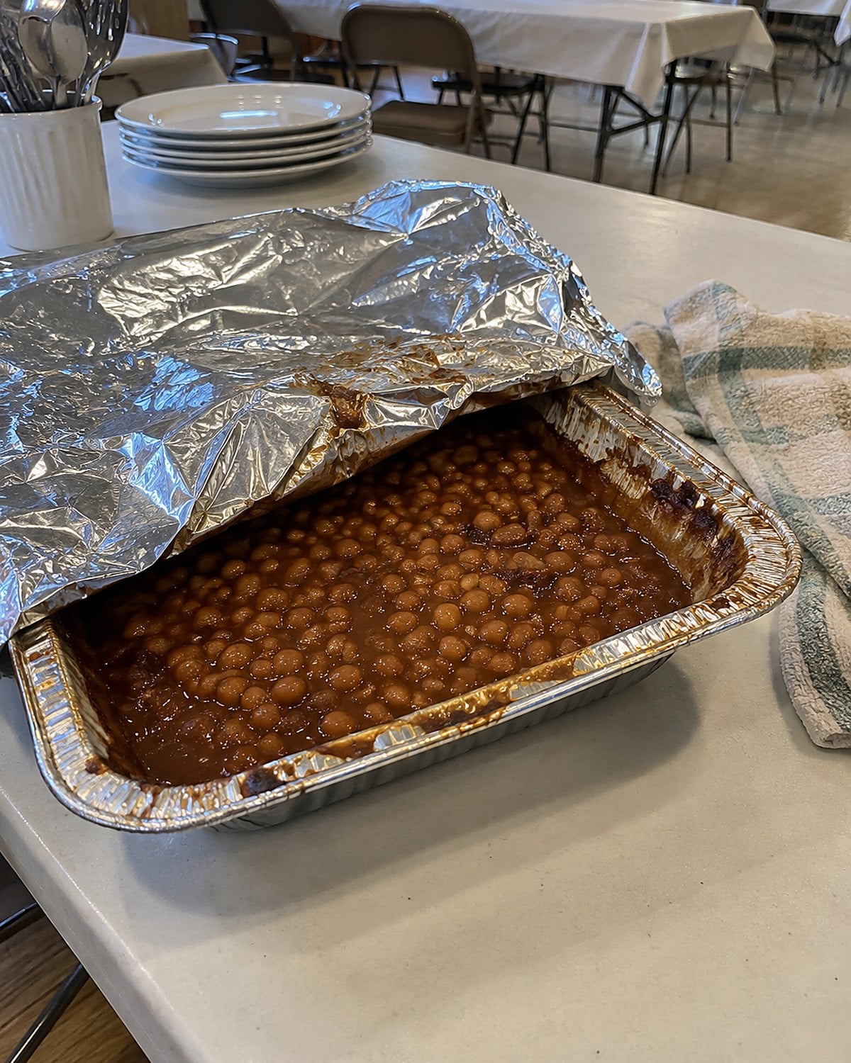 A foil-covered pan of baked beans partly uncovered on a church hall table.