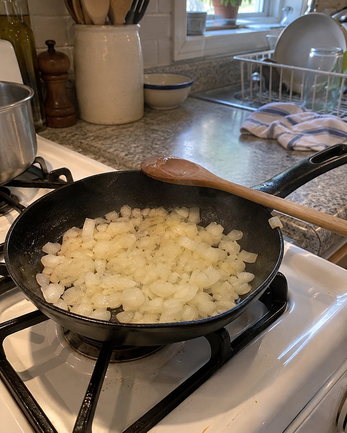 Chopped onion turning clear in a small skillet on a white stove, with a wooden spoon resting across the handle.