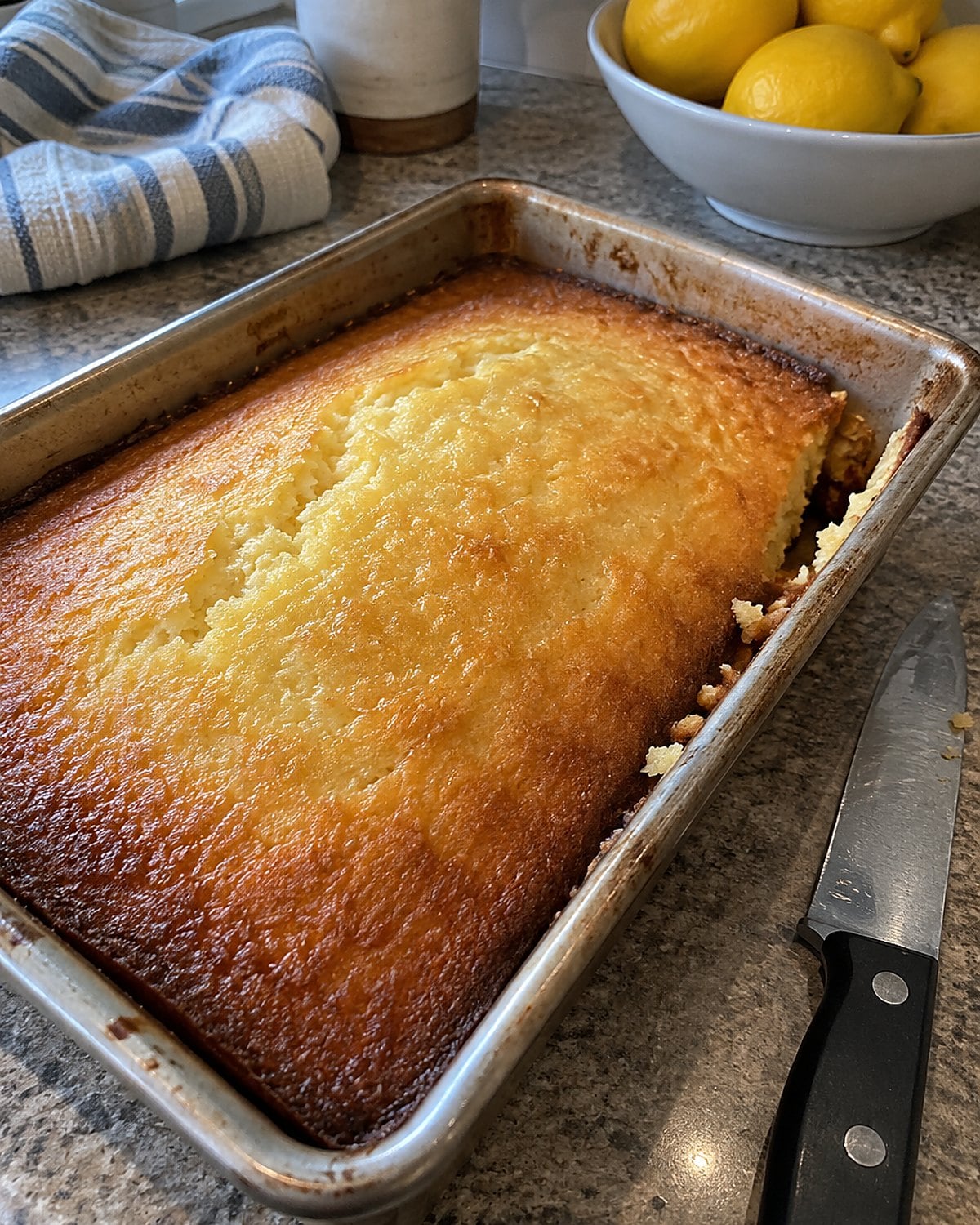 A lemon cake cooling in its pan on a counter beside a waiting knife.