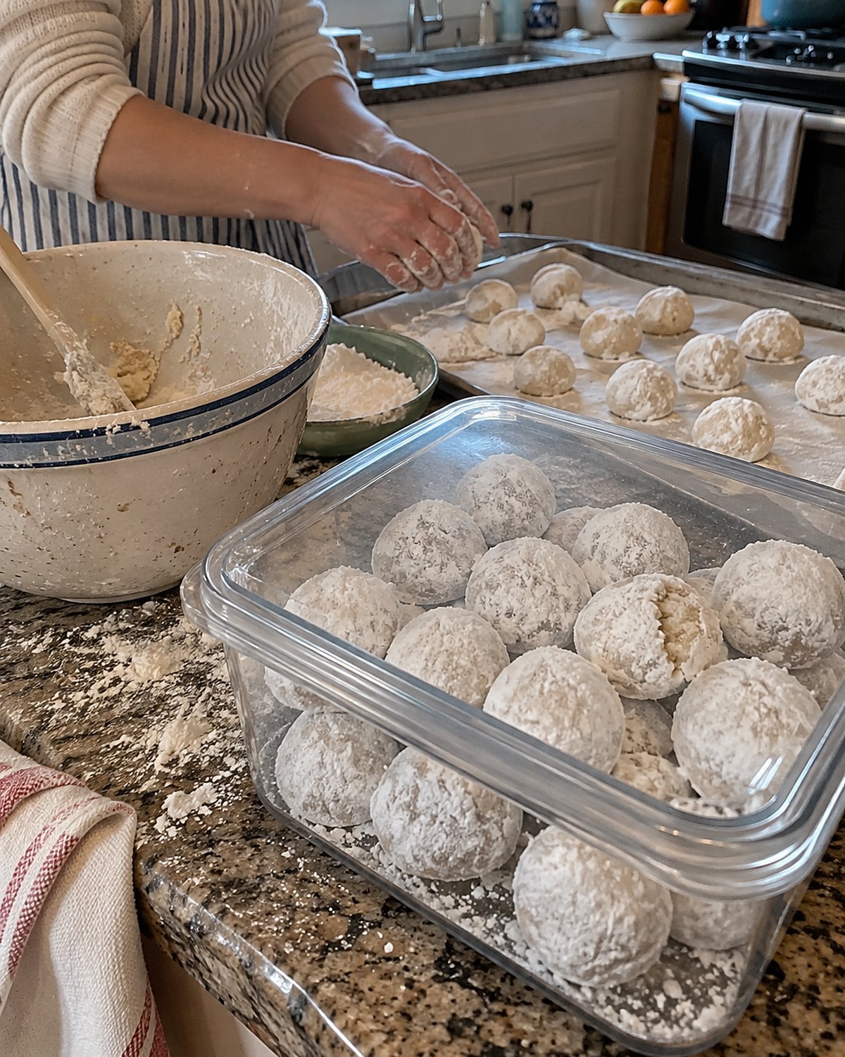 Powdered sugar snowball cookies in a covered container beside a flour-dusted mixing bowl.