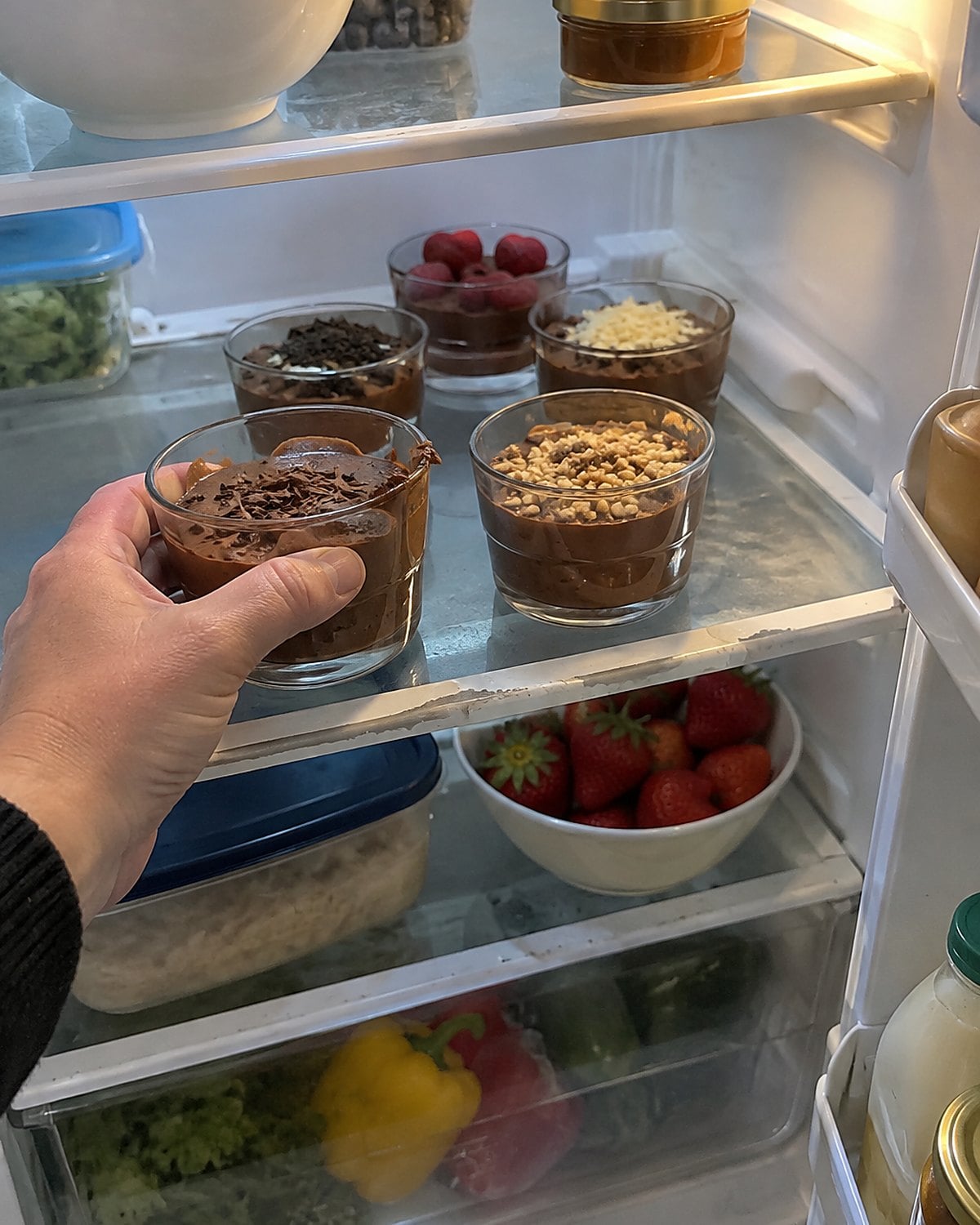 Small chocolate dessert cups with different toppings on separate refrigerator shelves.