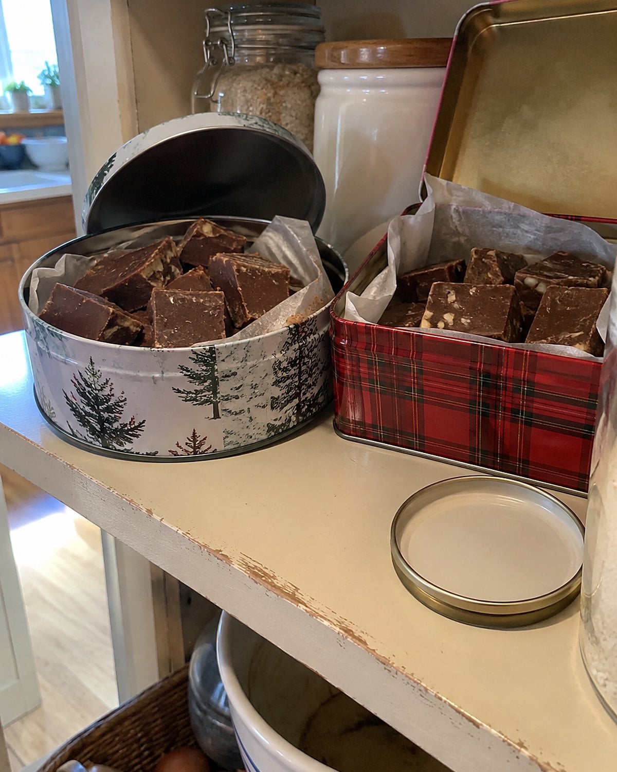 Two mismatched candy tins holding squares of fudge on a high pantry shelf.