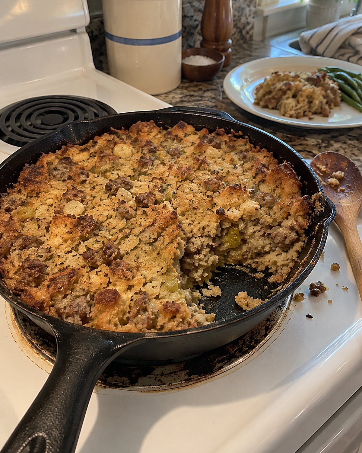 A cast-iron skillet of sausage dressing with one browned corner scooped out on a white stove.