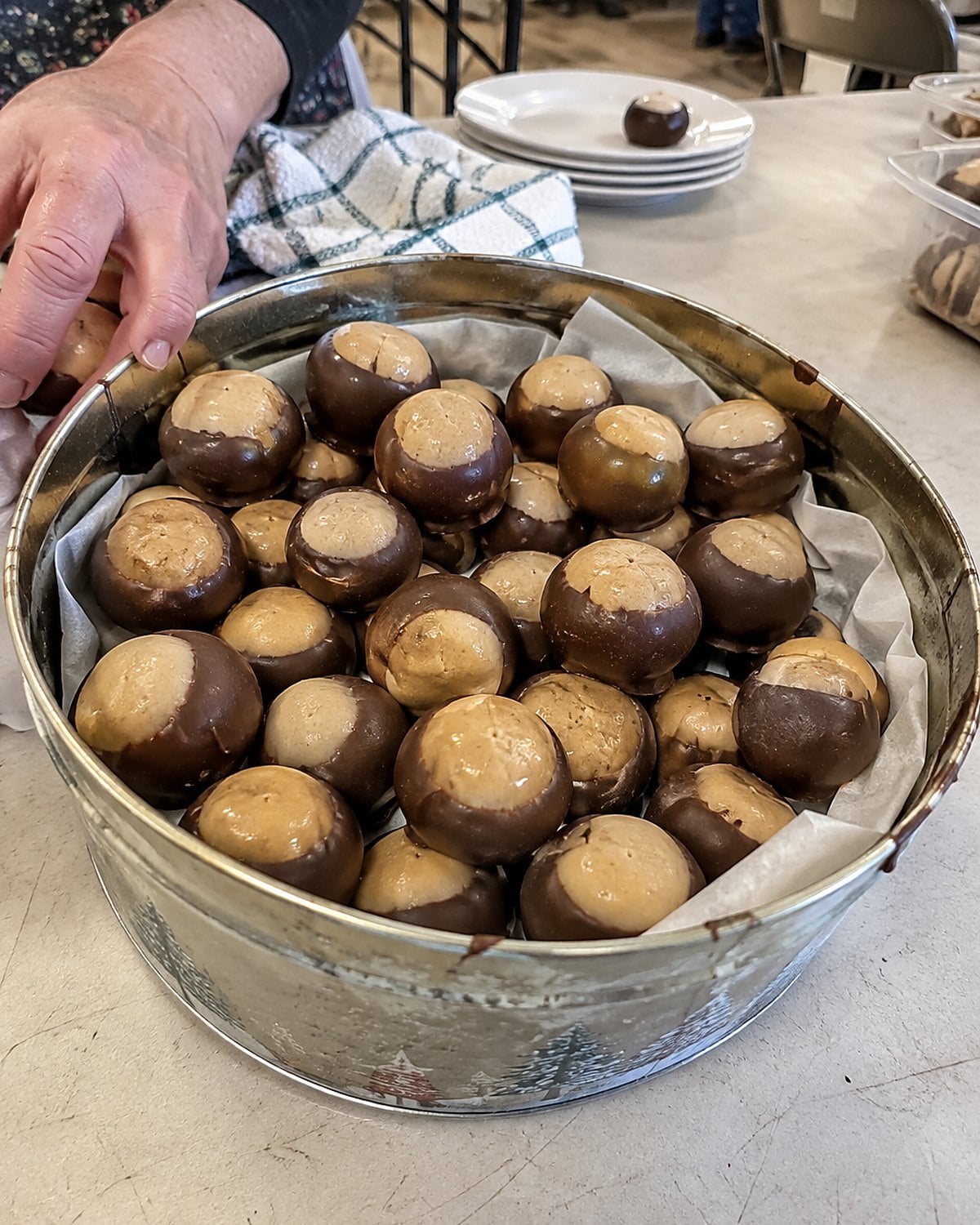 A dented cookie tin of chocolate-dipped buckeyes on a church hall table beside serving hands.