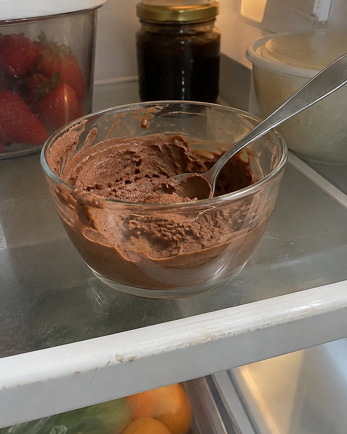 A spoon resting in a small glass bowl of chocolate mousse on a refrigerator shelf.