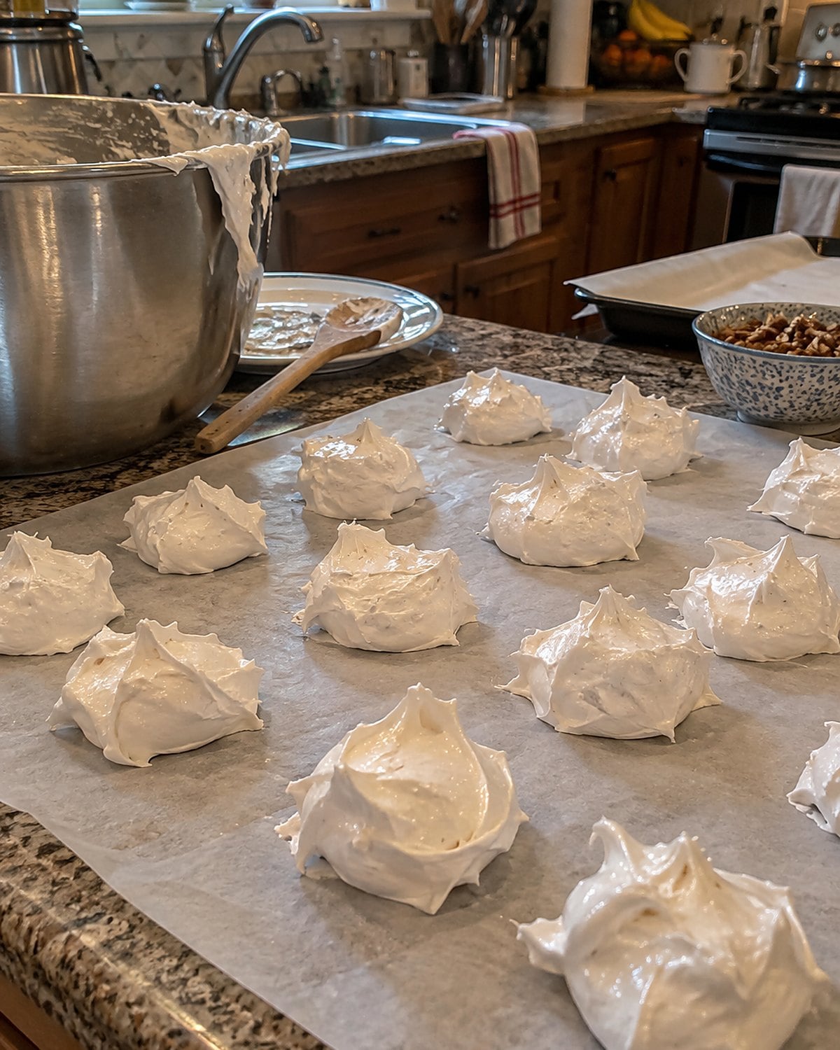 A mixer bowl beside uneven white divinity swirls cooling on wax paper across a crowded kitchen counter.