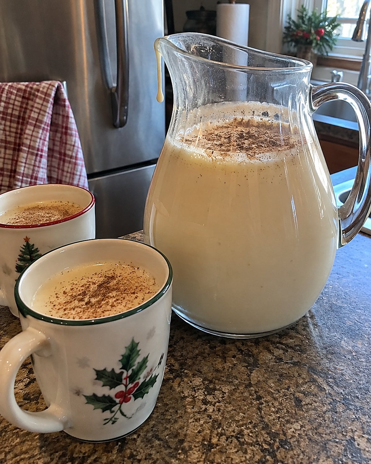 A glass pitcher of eggnog with nutmeg and two cups on a kitchen counter near the refrigerator.