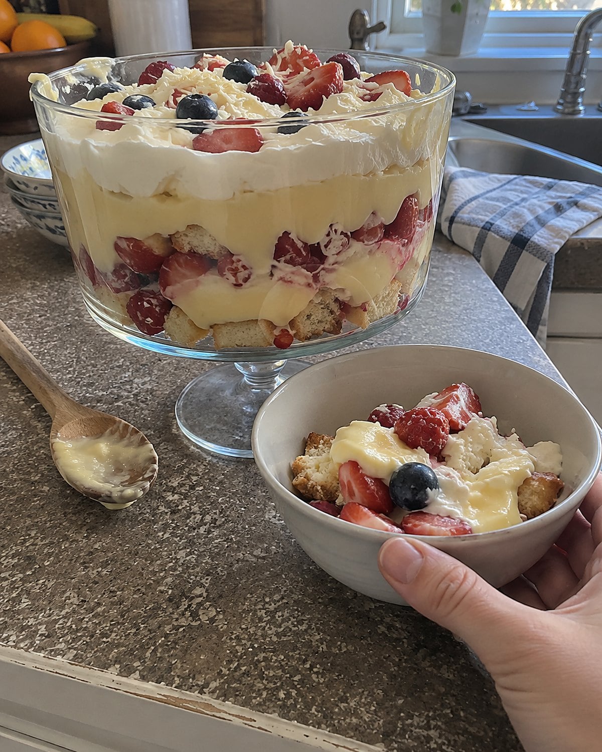 A large glass trifle bowl beside a smaller bowl of cake, fruit, and custard on the counter.