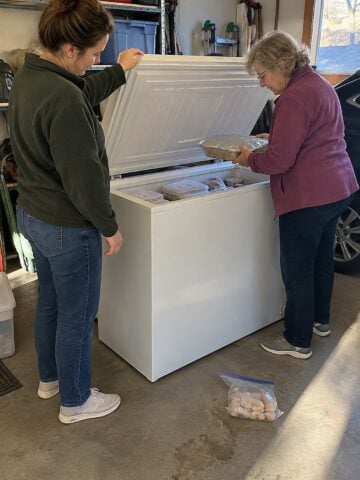 In a garage in late afternoon, a woman and her adult daughter lean into an open chest freezer as one lifts a foil pan and the other holds the lid.