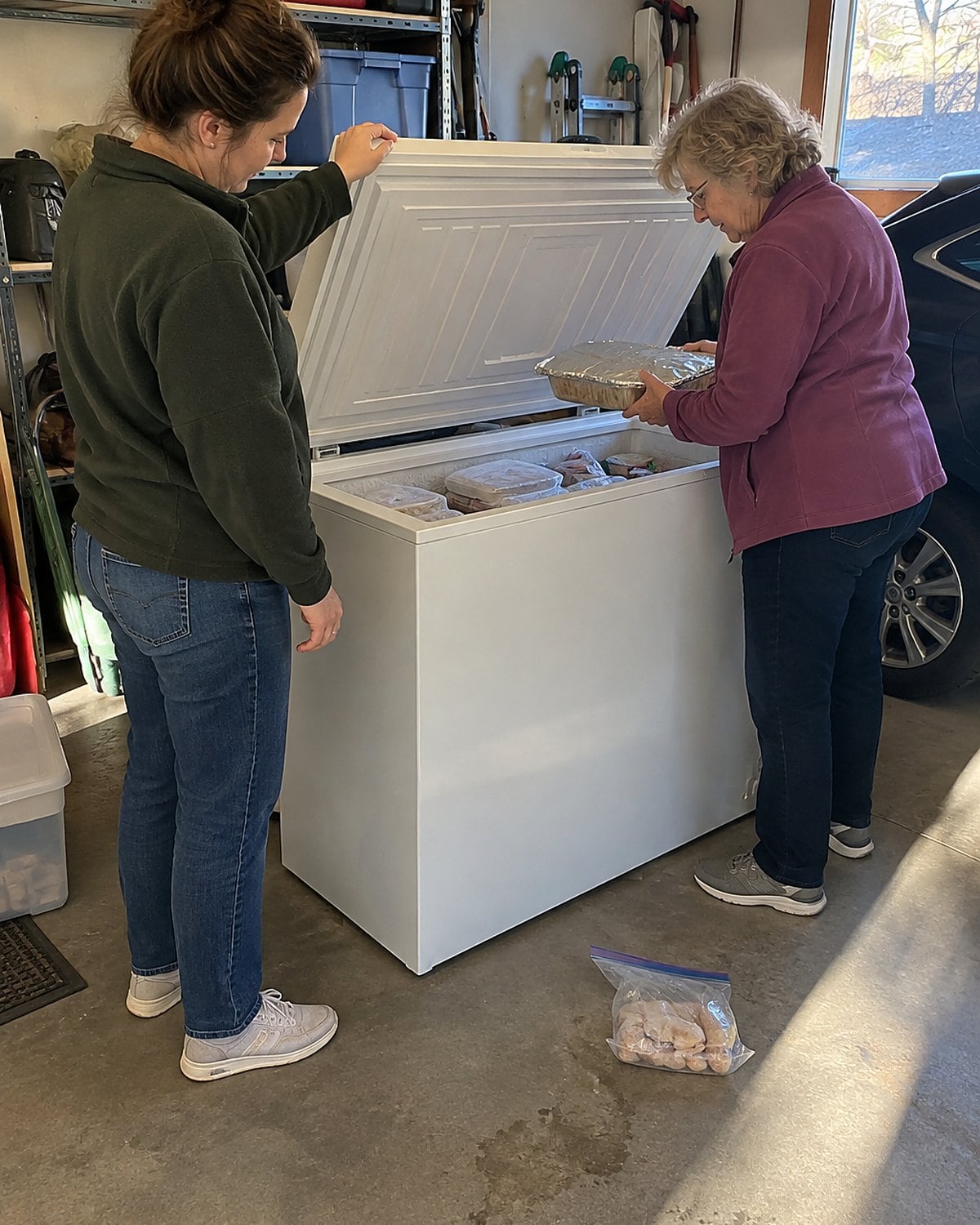 In a garage in late afternoon, a woman and her adult daughter lean into an open chest freezer as one lifts a foil pan and the other holds the lid.