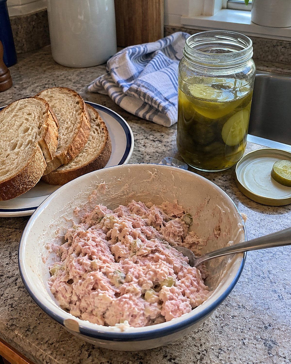A small bowl of ham salad sits beside sliced bread and a half-used jar of pickles.