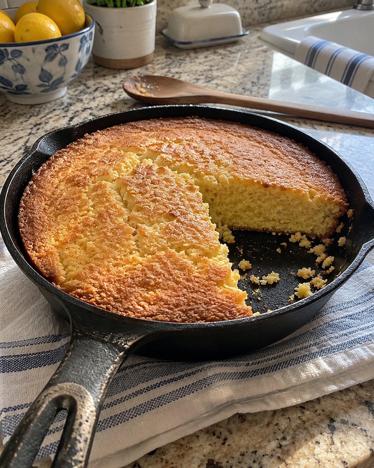 A cast-iron skillet of cornbread rests on a folded towel with a rough wedge cut from the edge.