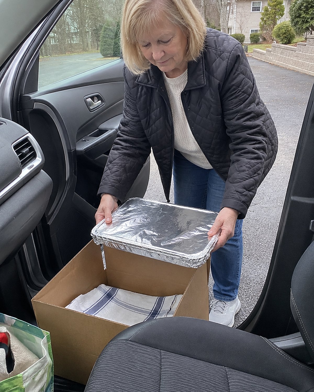Hands lower a covered foil pan into a sturdy box on the passenger-side floor of a parked car.