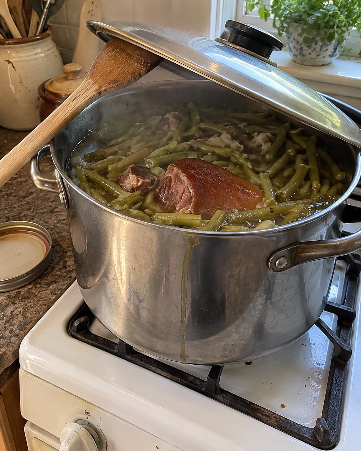 A dented pot of green beans and a ham hock rests on a stove with its lid propped on a spoon.