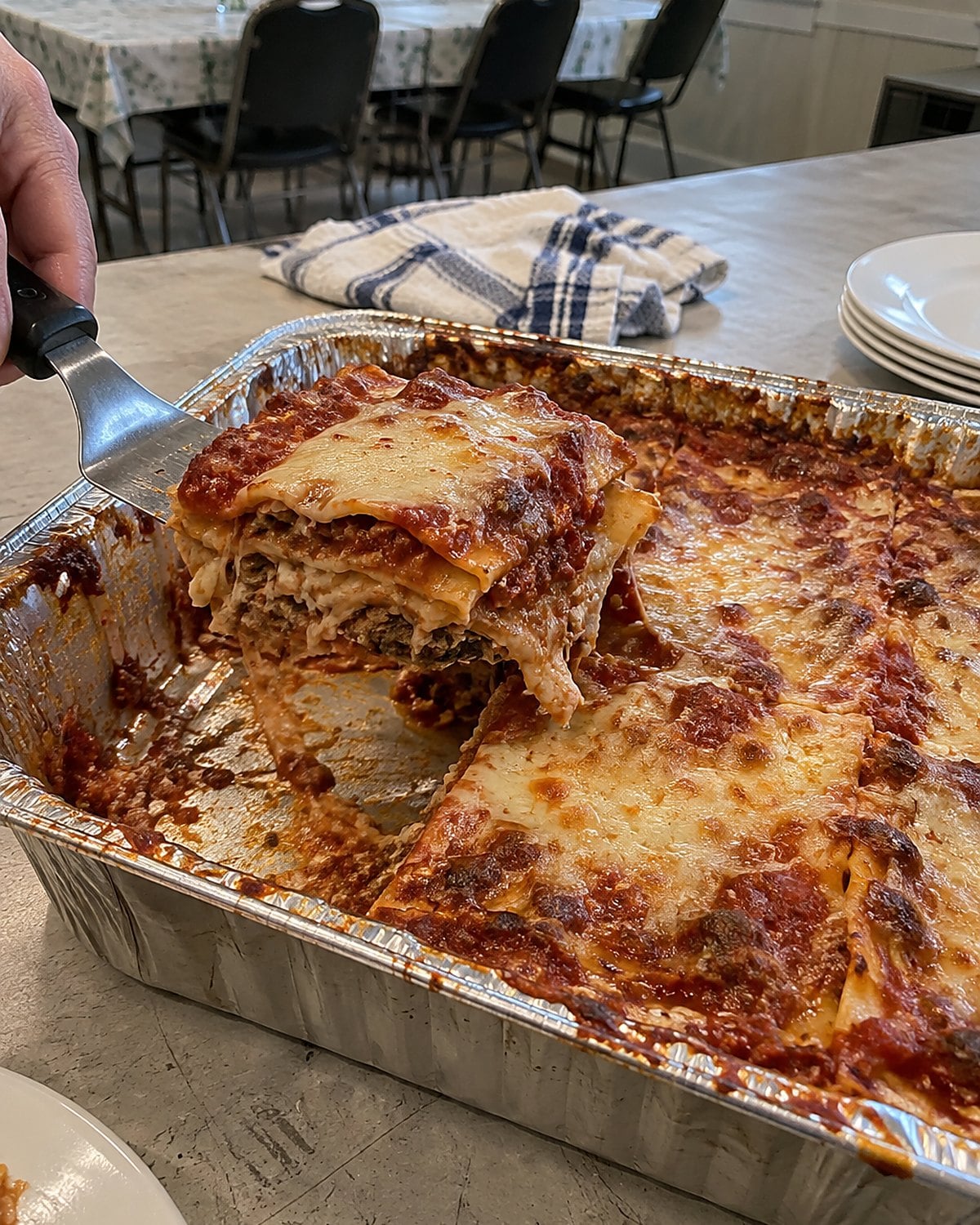 A broad spatula lifts a square of lasagna from a foil pan on a church hall serving table.