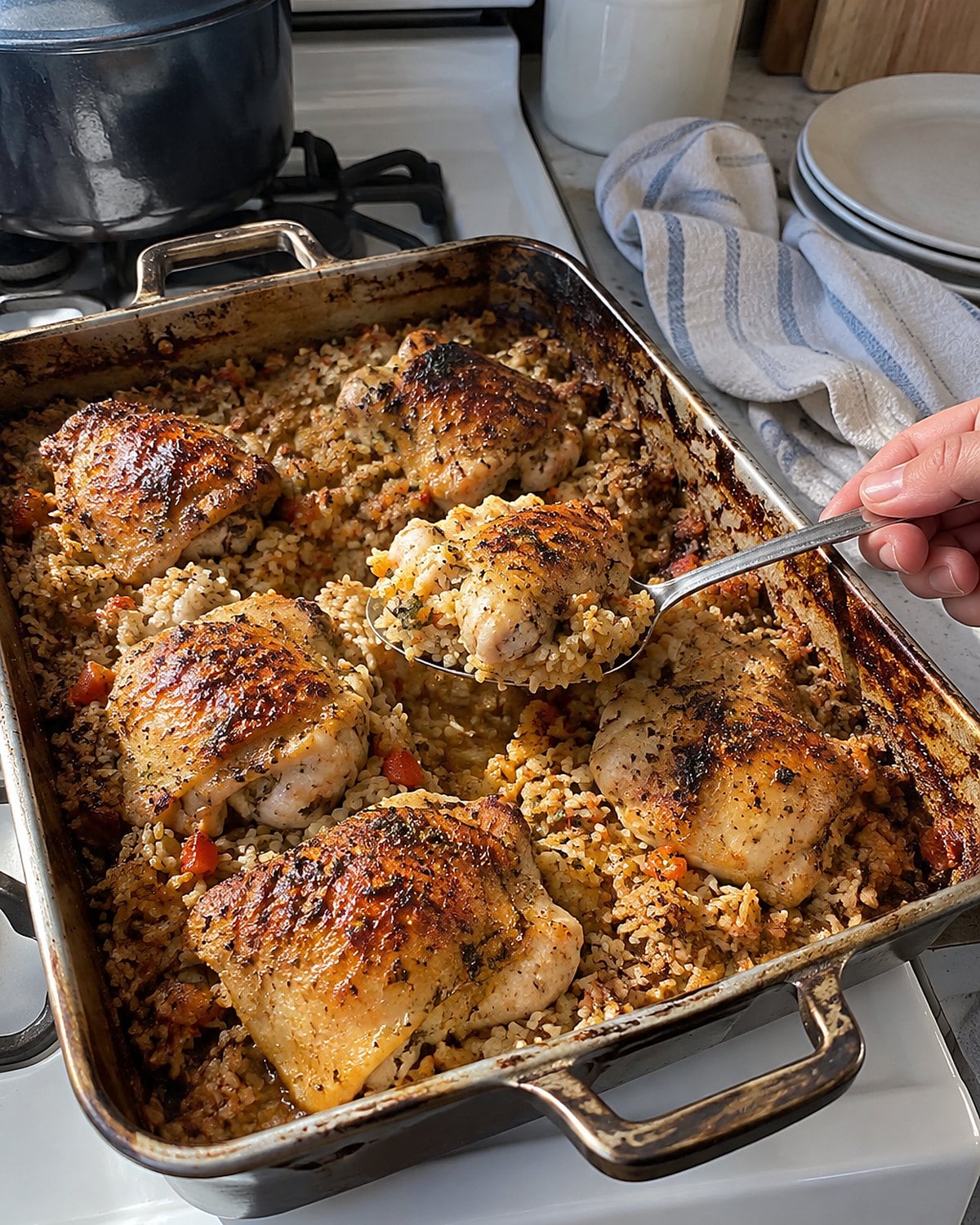 A spoon lifts chicken and rice from the center of a worn baking pan on a white stove, with grains clinging to its edge.
