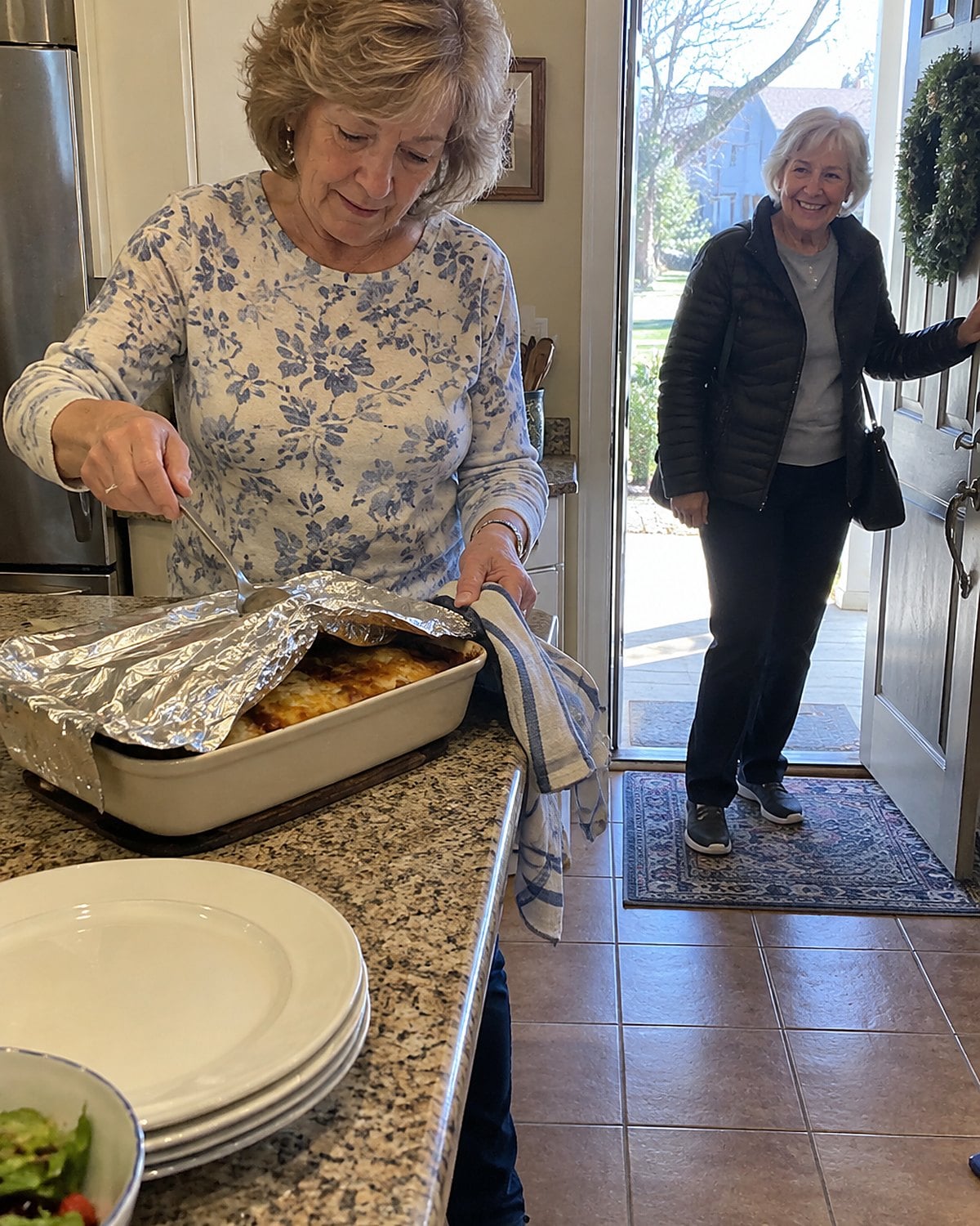 Hands tuck a serving spoon under the foil covering a casserole pan by the front door.