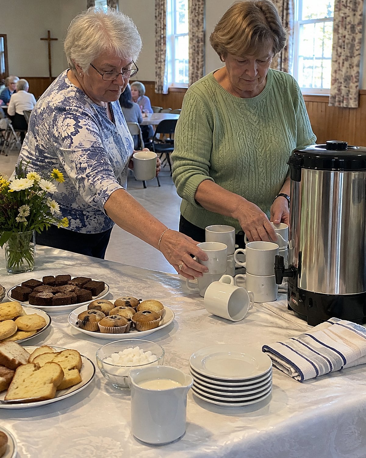 Hands set cups beside a coffee urn at the end of a church hall dessert table.