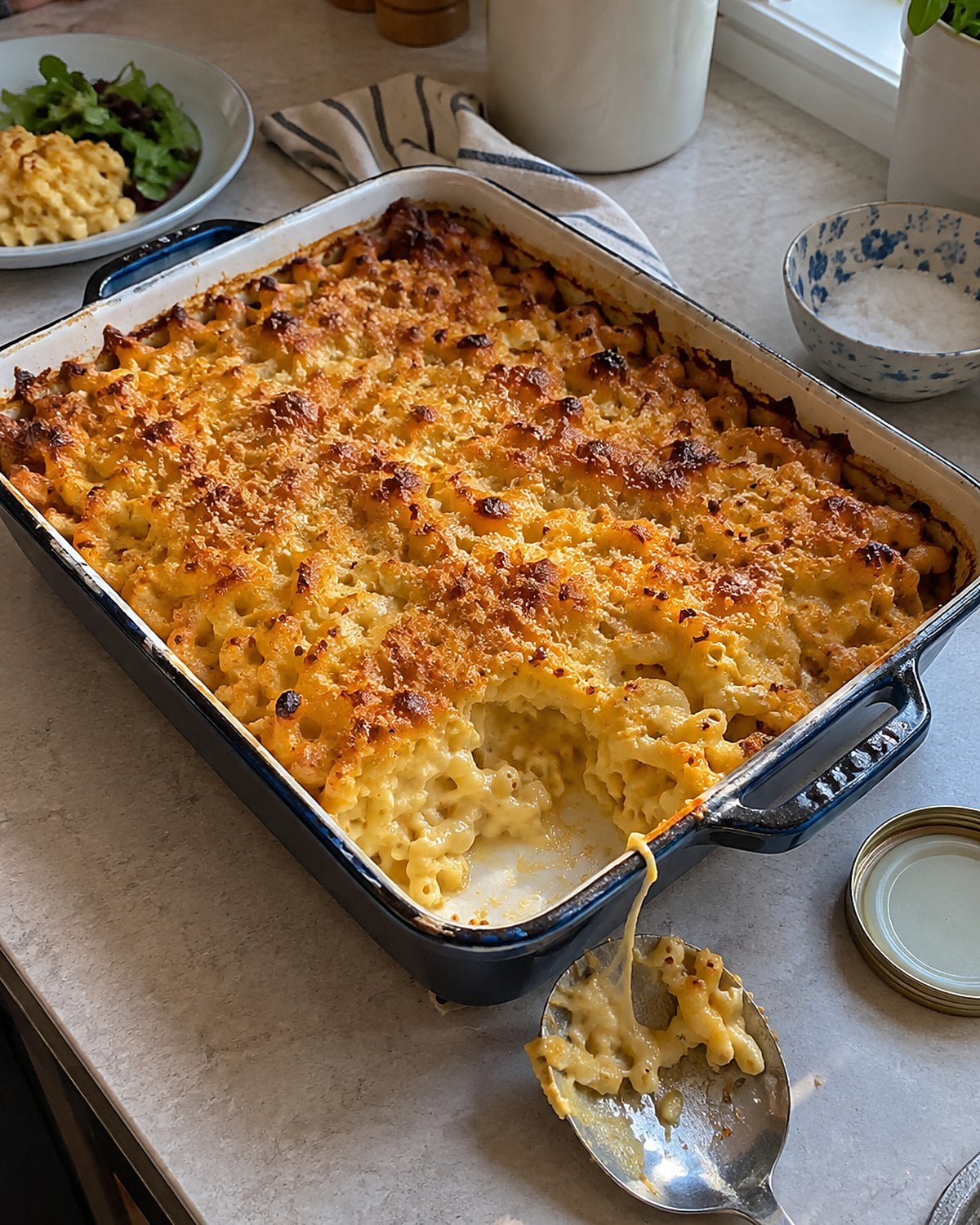 A baking dish of macaroni and cheese sits on the counter with browned edges and one spoonful taken.