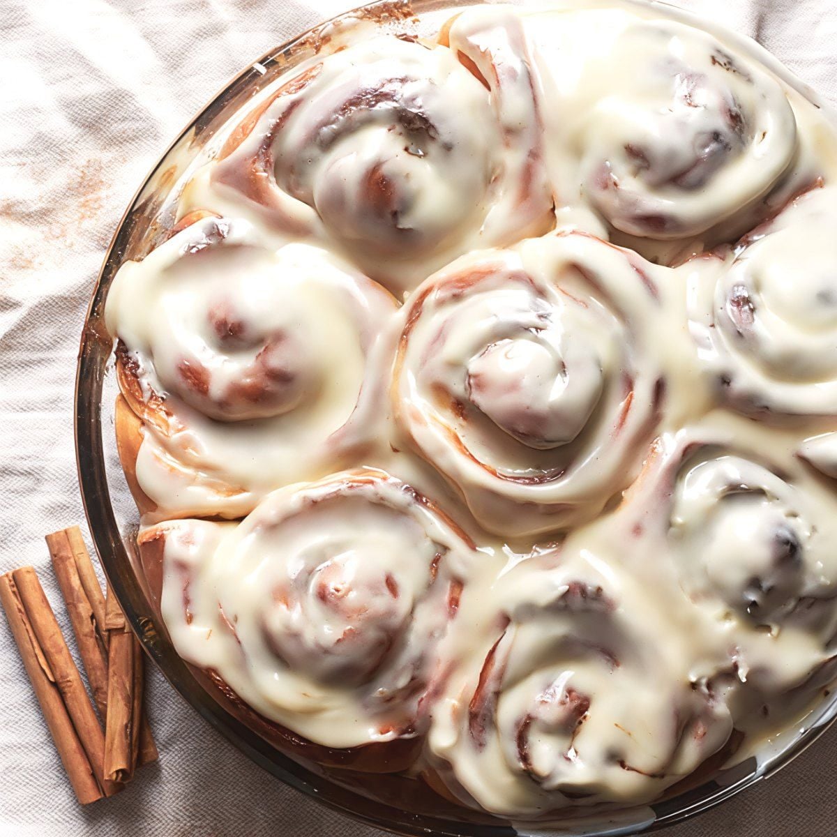 Freshly baked homemade cinnamon rolls topped with thick vanilla glaze served in a round baking dish next to cinnamon sticks for Thanksgiving breakfast ideas.