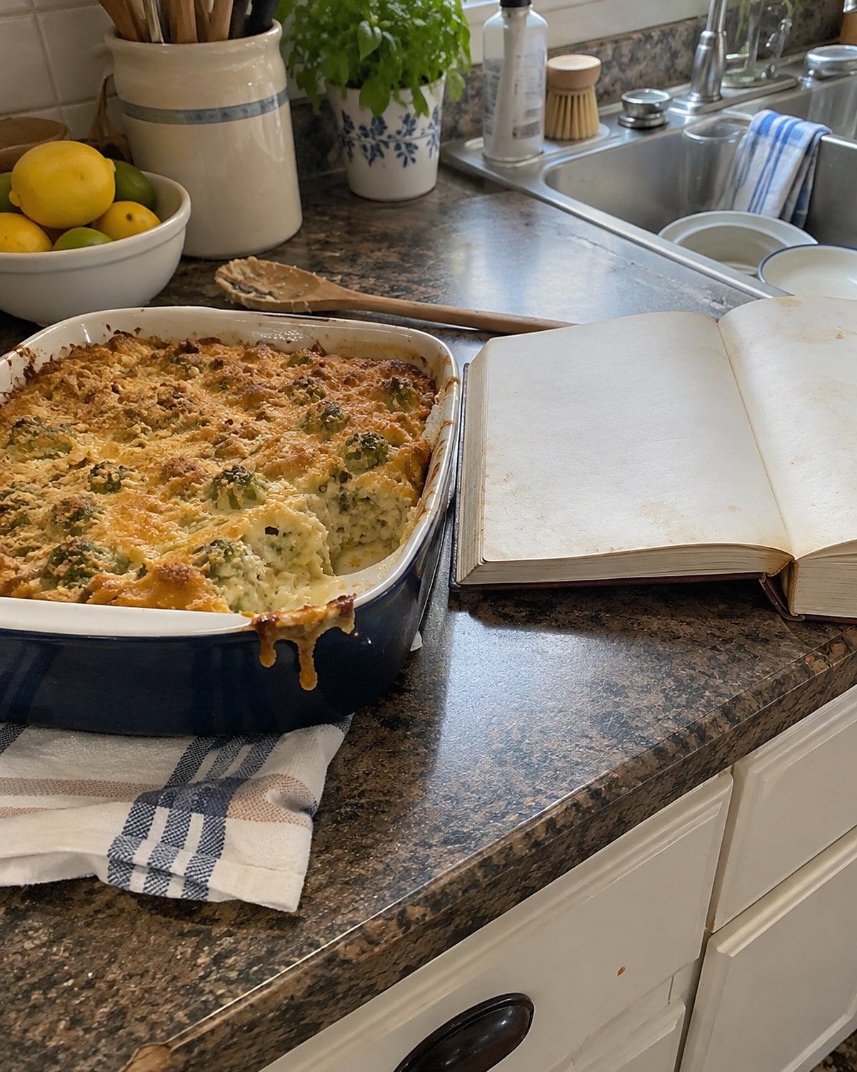 A stained church cookbook lies open beside a smaller casserole dish on a kitchen counter.
