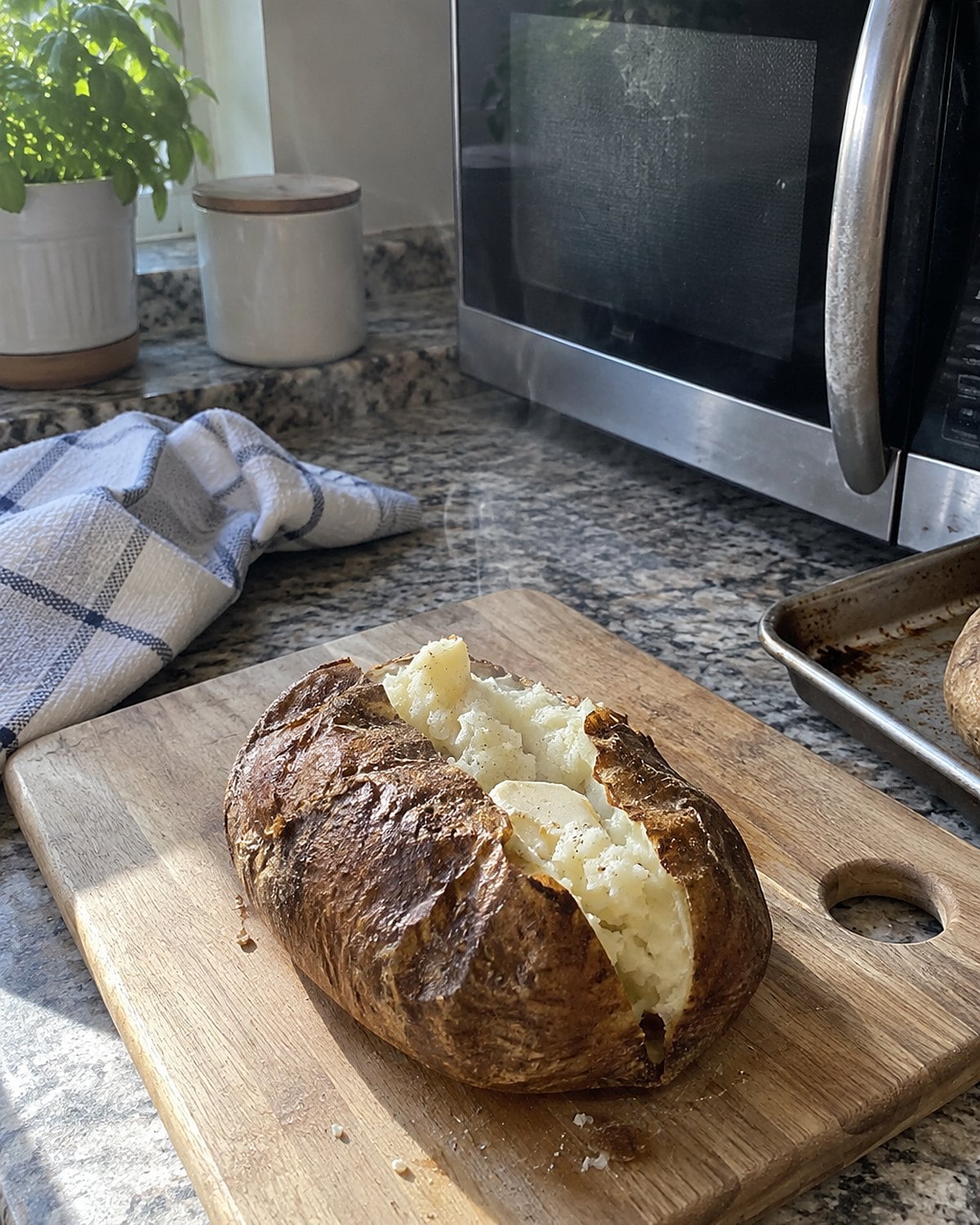 One baked potato rests on a cutting board beside a microwave and a small baking sheet.