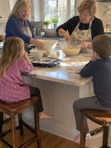 Two women friends work at a kitchen island mid-afternoon while grandchildren sit on stools nearby, one friend reaching for a mixing bowl as the other stirs, nobody posing.
