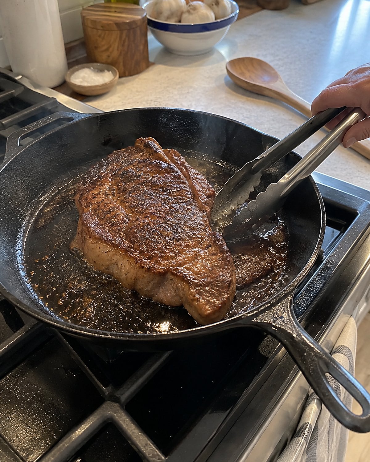 Tongs beside a steak with a browned crust in a cast-iron skillet.