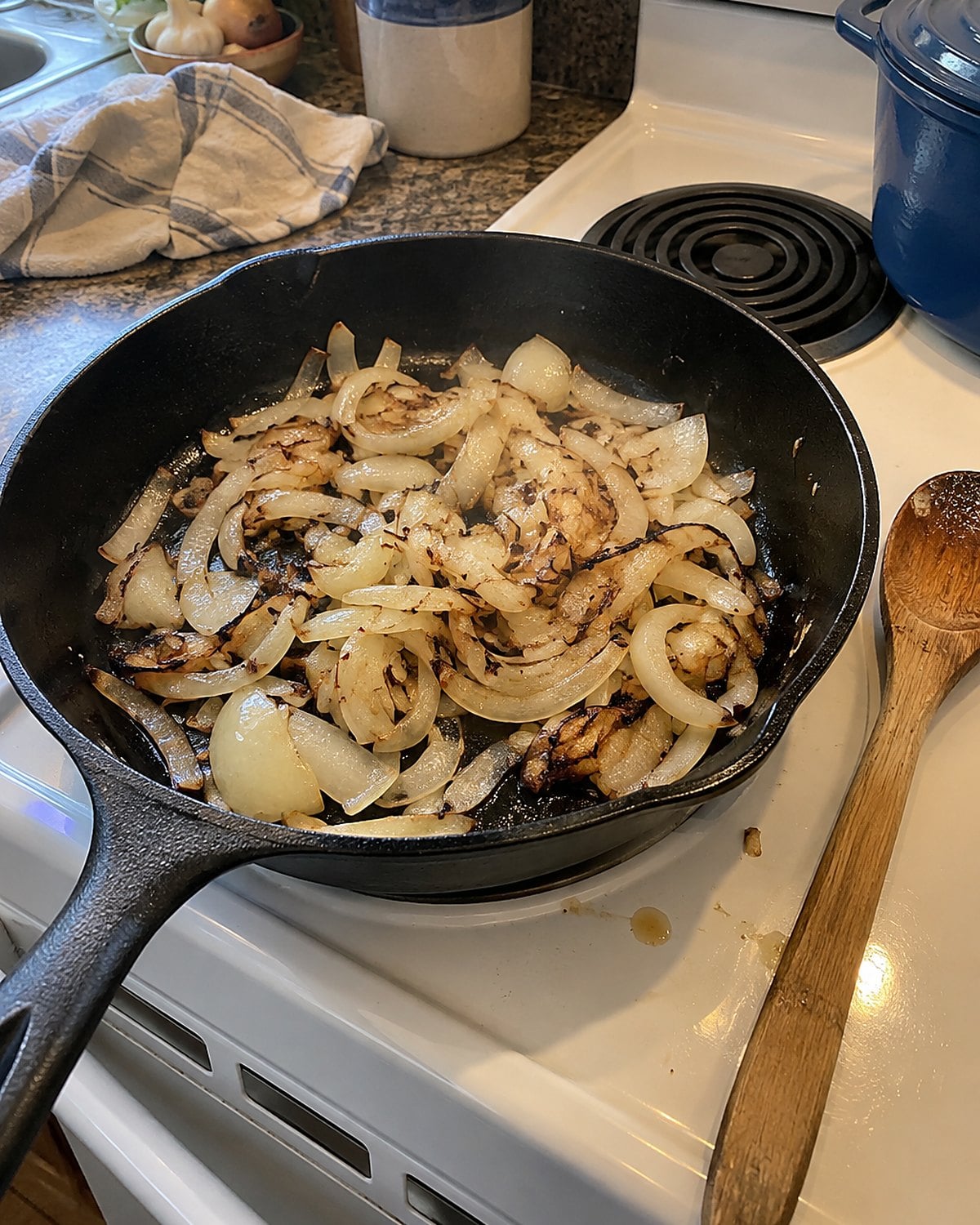 A wooden spoon resting beside a skillet of partly browned onions on a white stove.