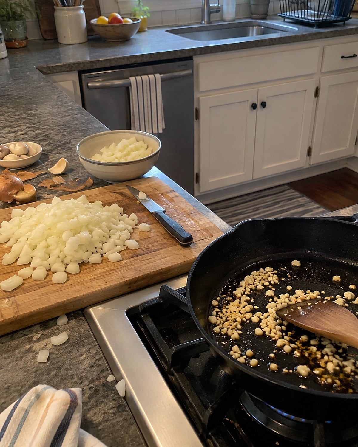 Chopped onions waiting on a cutting board beside garlic browning in a skillet.