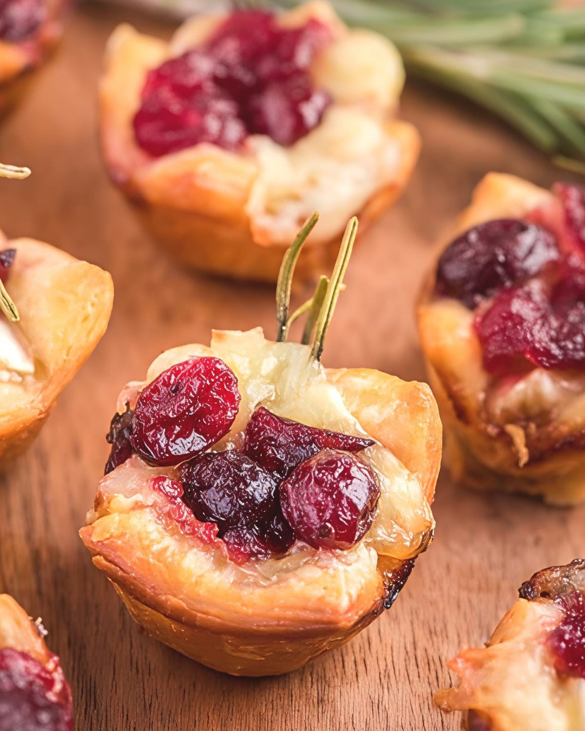 Warm, golden puff pastry Cranberry Brie Bites filled with melted brie cheese, sweet cranberry sauce, and fresh rosemary sprigs on a wooden board, ideal as quick and festive last minute potluck ideas.