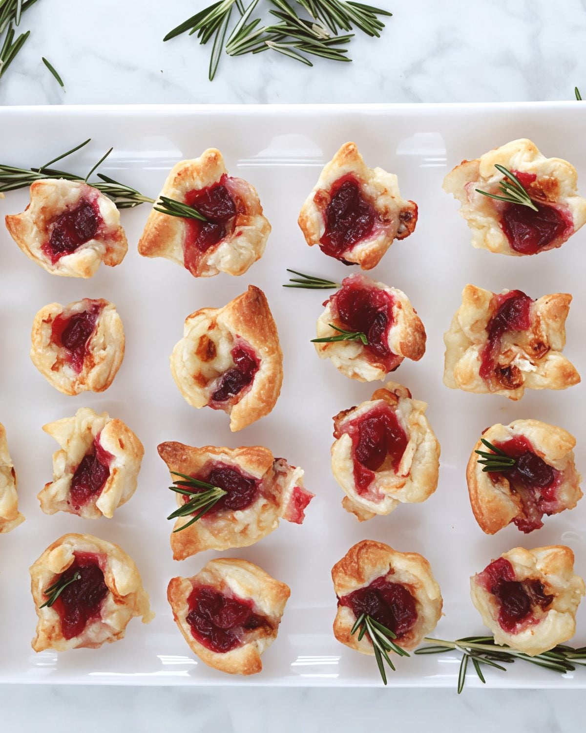 Golden puff pastry cranberry brie bites topped with chopped pecans and fresh rosemary sprigs, perfect for Thanksgiving side dishes for a crowd.