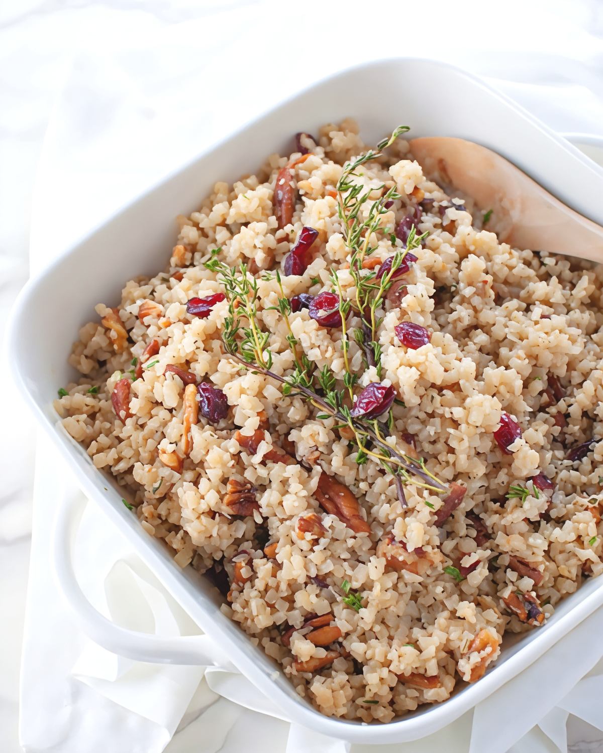 A white serving dish filled with cauliflower rice stuffing mixed with dried cranberries, crunchy pecans, and fresh thyme sprigs makes a delicious addition to keto thanksgiving sides.