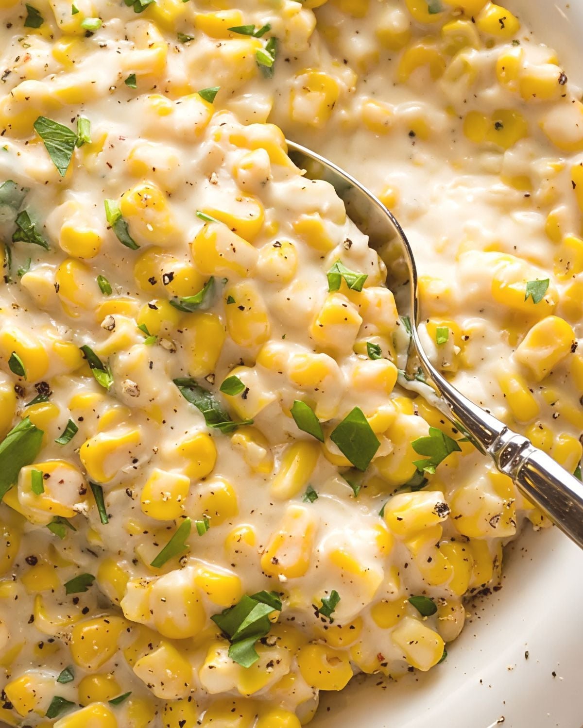 A close-up view of rich, creamy corn garnished with fresh chopped herbs and cracked black pepper, served with a silver spoon.