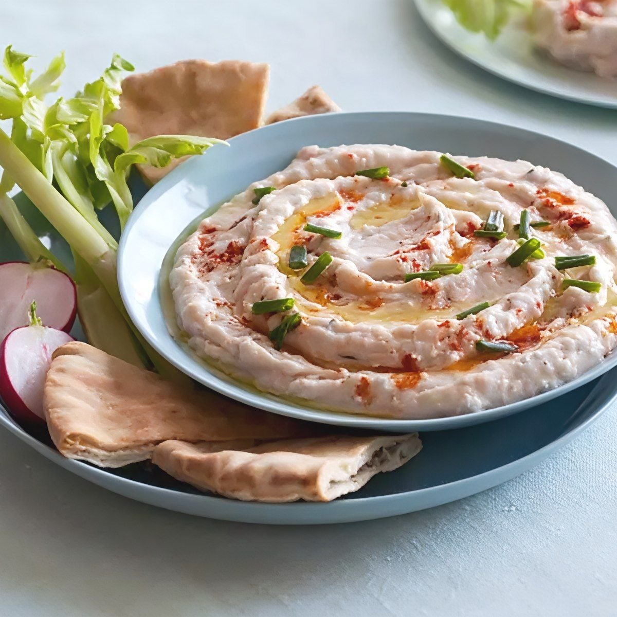 Lighter creamy white bean dip swirled with olive oil, paprika, and chopped chives, served with pita bread, celery sticks, and sliced radishes as a healthy Thanksgiving appetizer.