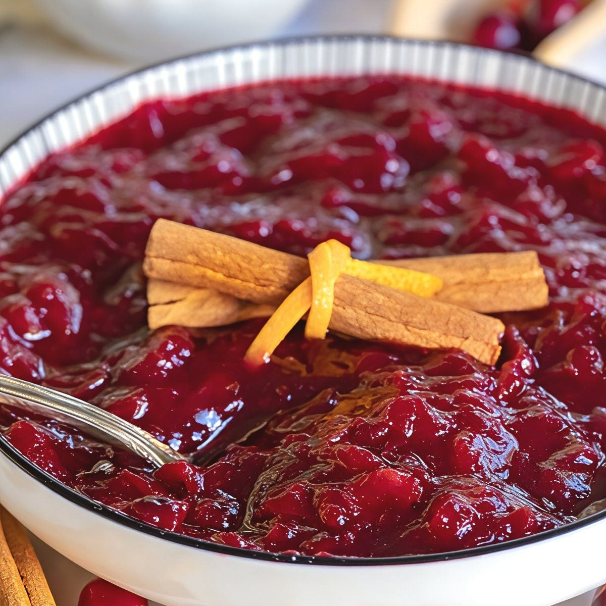A white bowl of vibrant homemade slow cooker cranberry sauce garnished with cinnamon sticks and orange peel, featured as one of the best crockpot thanksgiving sides.