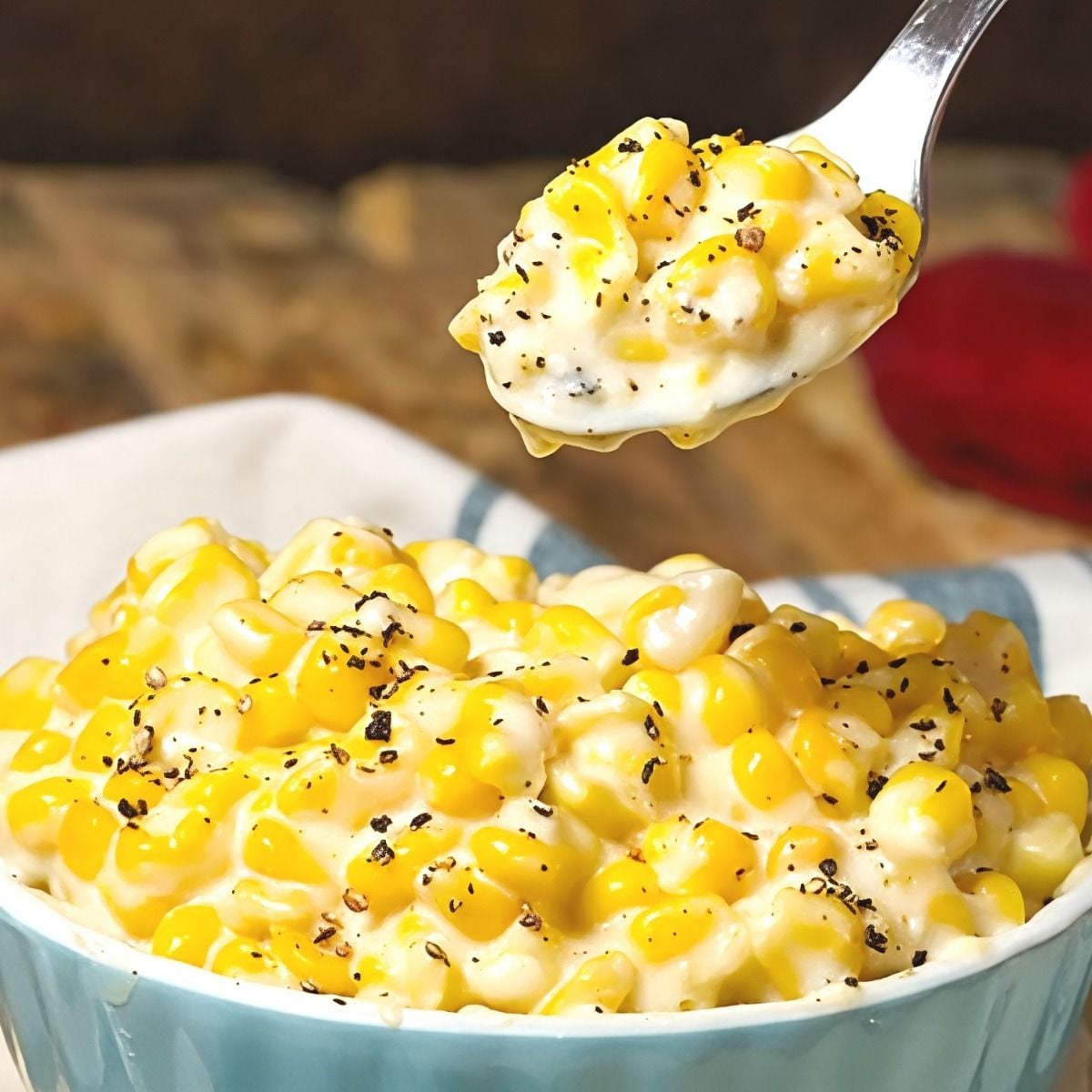 A spoon lifting a bite of rich and creamy slow-cooked corn seasoned with cracked black pepper from a blue bowl, featured as one of the best crockpot thanksgiving sides.