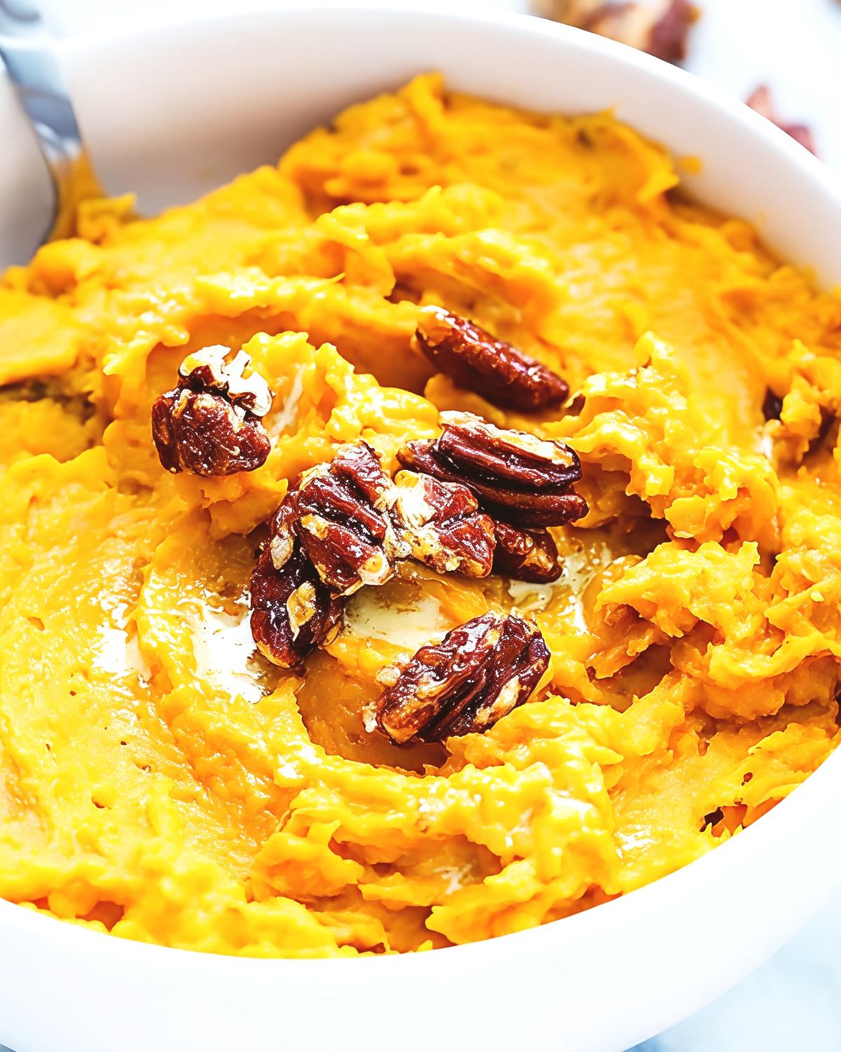 A white bowl filled with creamy slow-cooked mashed sweet potatoes topped with crunchy pecan halves and melted butter, perfect for crockpot thanksgiving sides.