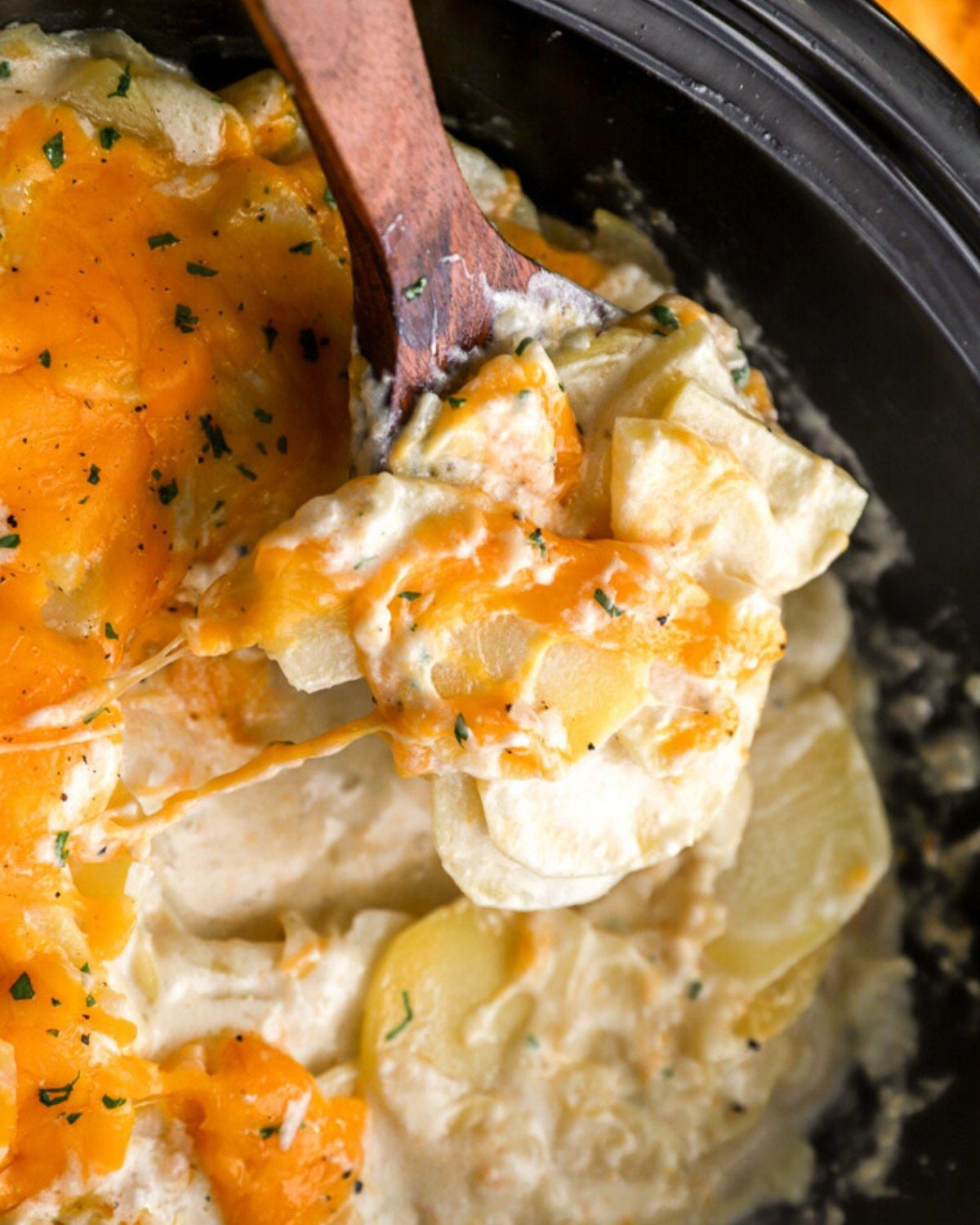 A wooden spoon scooping creamy cheesy scalloped potatoes directly from a slow cooker, featured as one of the best crockpot thanksgiving sides.