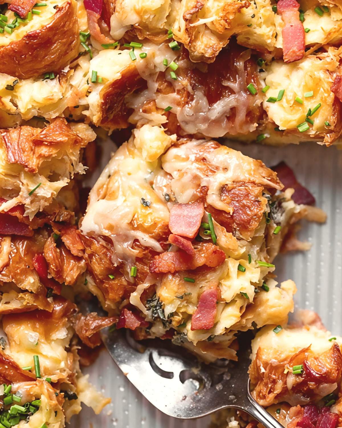 A close-up of a savory croissant breakfast casserole with bacon and gruyere being scooped with a spoon as Thanksgiving breakfast ideas.