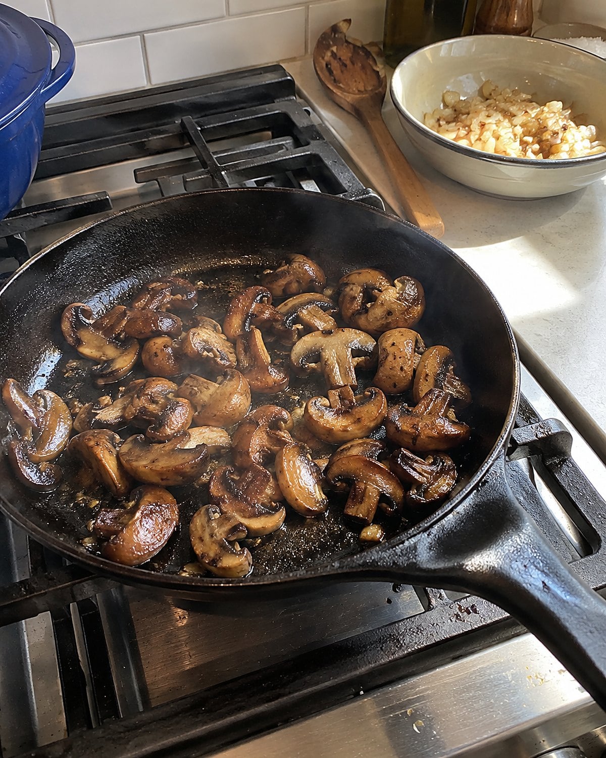 Mushrooms brown in an uncrowded skillet while cooked onions wait in a bowl by the stove.