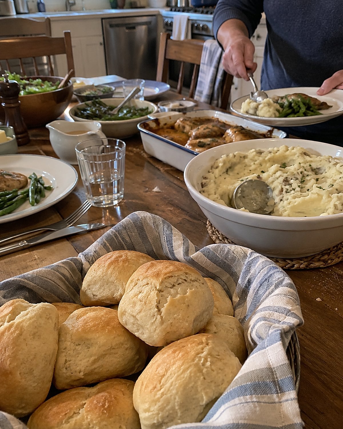 A basket of uneven homemade rolls sits beside the main dishes on a set dinner table.