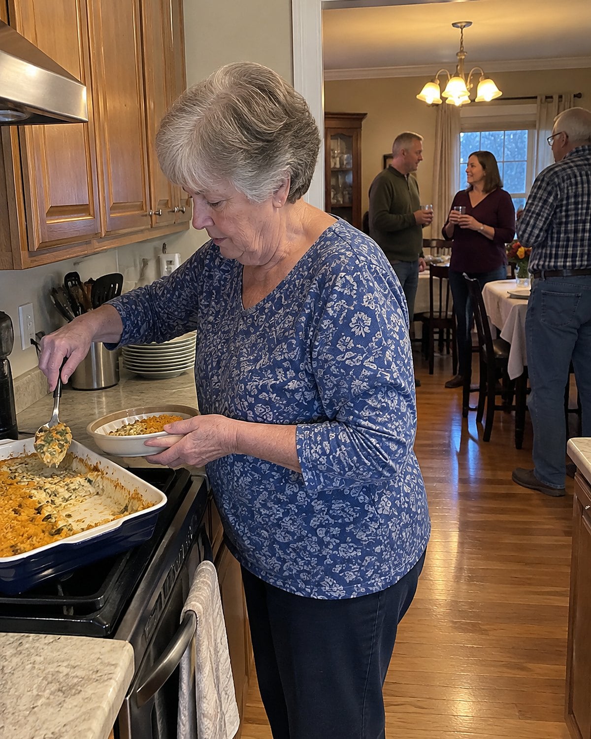 An older woman spoons casserole into a shallow container beside the stove while guests talk in the next room.