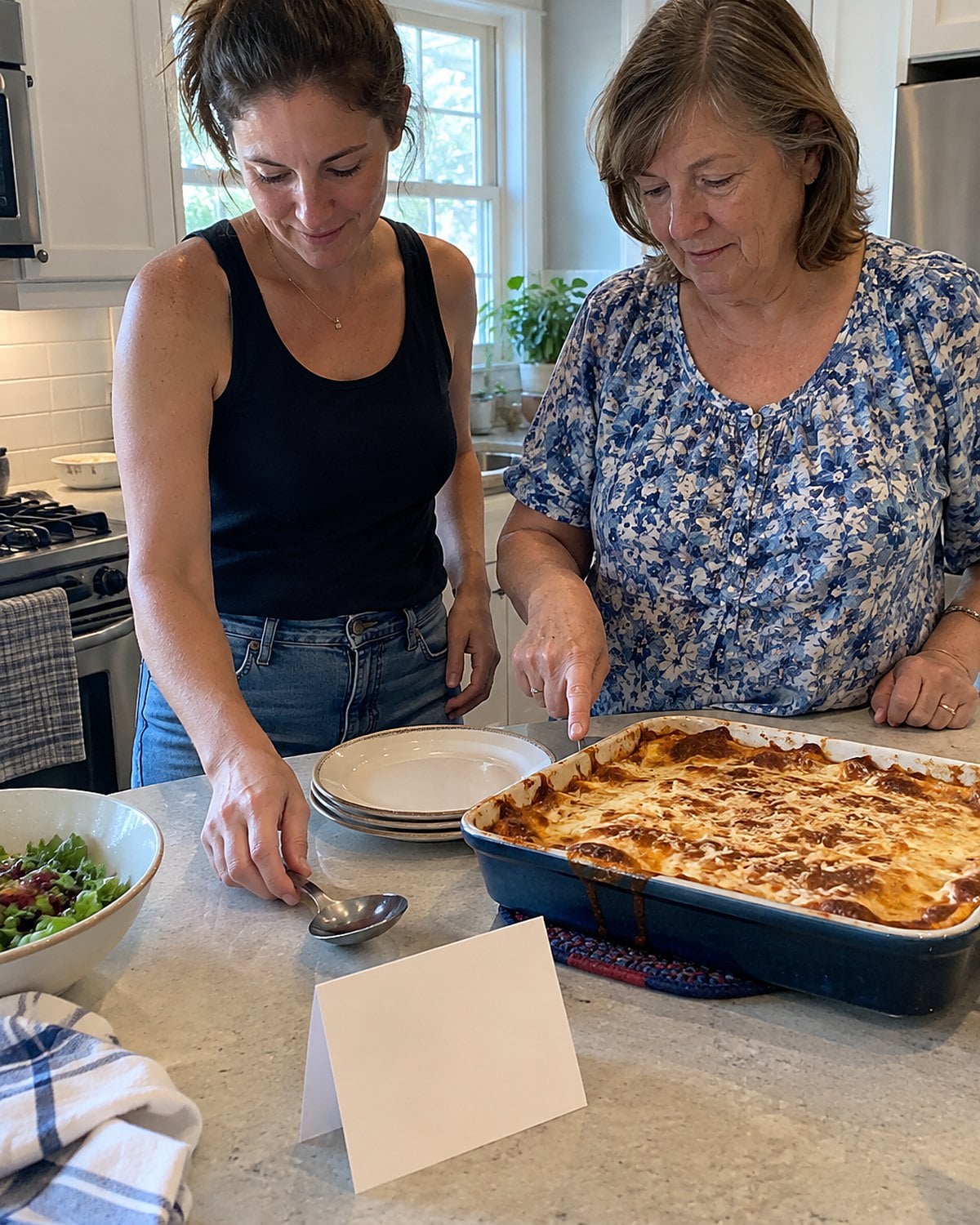 Two women stand at a kitchen counter beside an old recipe card and a casserole dish.